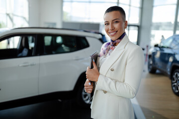 Saleswoman in car showroom holding a laptop computer and selling cars.