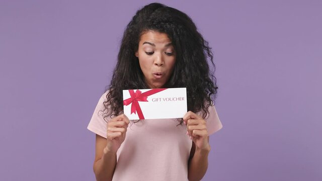 Surprised happy young african woman 20s years old wears pink T-shirt pointing finger on gift certificate voucher for store doing winner gesture isolated on pastel violet purple color background studio