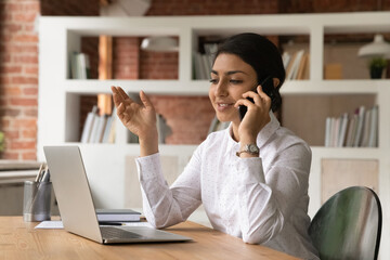 Successful happy young indian mixed race business woman holding cellphone call conversation, negotiating with client distantly working on computer in modern office, professional multitasking concept.