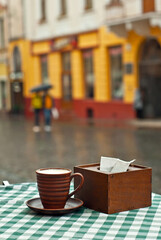 Coffee in a clay mug on a checkered tablecloth. Terrace of a street cafe on a background of blurred street. Sharpness on the mug. People with umbrellas on the background.