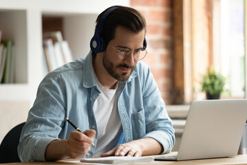 Happy focused young man in eyeglasses wearing wireless headphones, watching educational lecture on computer, studying on online courses distantly, improving professional knowledge, writing notes. - obrazy, fototapety, plakaty