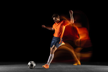 Plakat Young caucasian male football or soccer player kicking ball for the goal in mixed light on dark background. Concept of healthy lifestyle, professional sport, hobby.