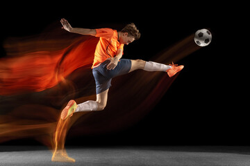 Young caucasian male football or soccer player kicking ball for the goal in mixed light on dark...