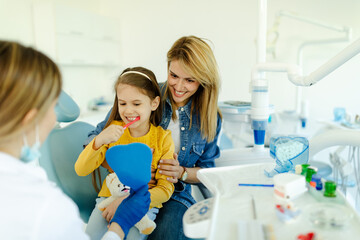 Girl looking in the mirror and brush her teeth after dental procedure while mother sitting near her.
