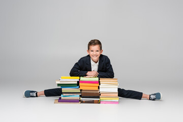 flexible schoolboy doing split near pile of books on grey.