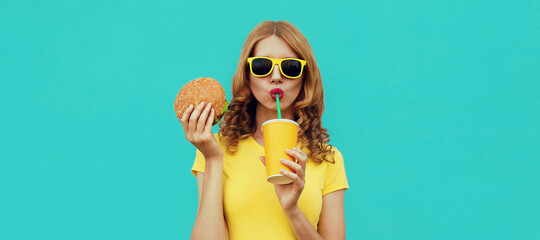 Portrait close up of young woman with fast food, burger and cup of juice wearing a yellow t-shirt,...