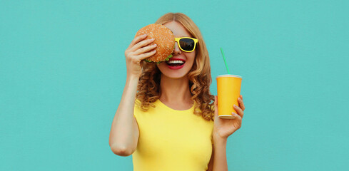 Portrait of happy smiling young woman with fast food, burger and cup of juice wearing a yellow...