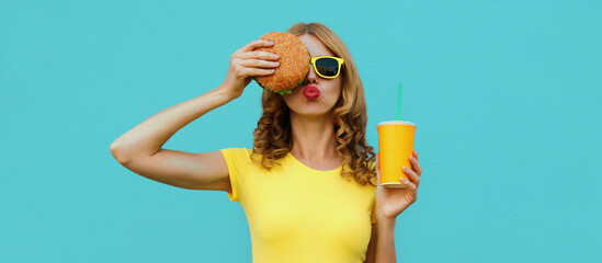 Portrait close up of young woman with fast food, burger and cup of juice wearing a yellow t-shirt,...