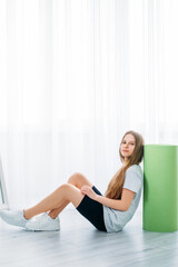 Obraz na płótnie Canvas Kids home sport. Fitness indoors. Active children. Girl in sportswear sitting on floor with green yoga mat on light white window copy space background.