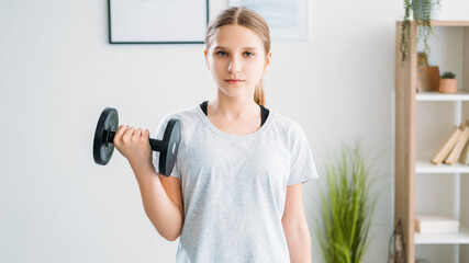 Home fitness. Active kids. Morning training. Strength wellbeing. Athletic girl in activewear doing physical exercise with dumbbell for arm biceps muscles in light room interior.