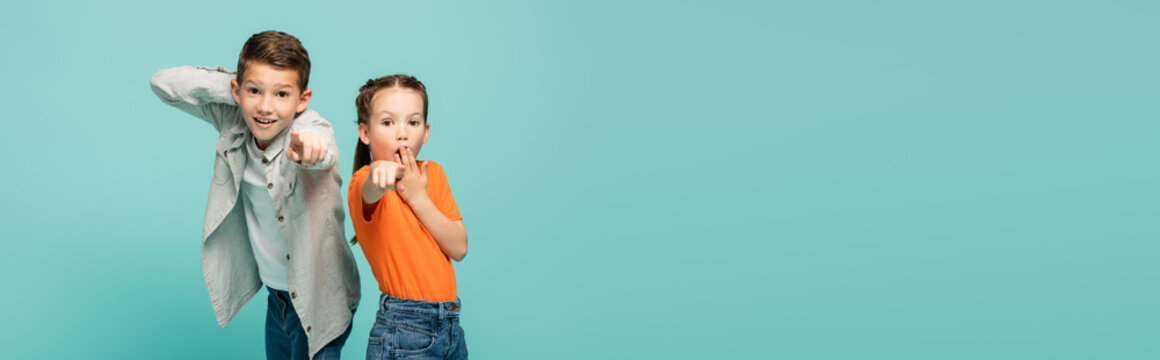 shocked girl in orange t-shirt covering mouth near boy pointing at camera isolated on blue, banner.