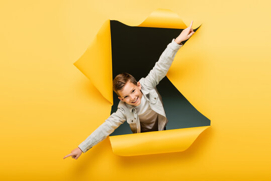 Joyful Boy With Outstretched Hands Looking At Camera Through Ripped Hole On Yellow Background.