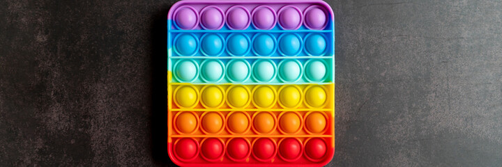 kids anti stress sensory pop it or simple dimple fidget push toy on a black table or floor background. square popit rainbow hue bright color, trend 2021 year. top view, flat lay. banner
