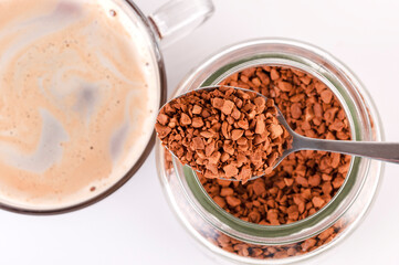 The spoon contains dry granules of instant coffee. A coffee drink is poured into the cup.