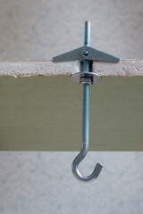 Folding metal dowel hook with spring-loaded wings installed in waterproof sheet of drywall against the background of blurred interior wall, view of section of gypsum fiber in the place of fixation.
