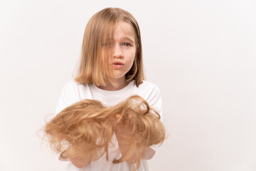 upset little girl holds in hands cropped hair after cutting on a white background. means to care for children's hair. Beauty salon for kids.