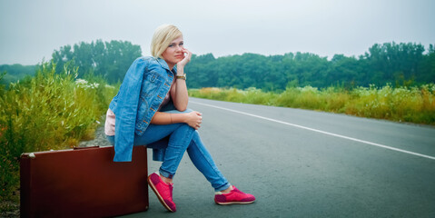 Portrait of beautiful blond female traveler siting on suitcase, looking away on road and waiting...