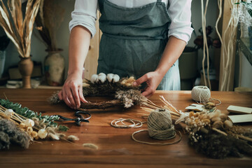 Hands of Female Florist Making Wreath with Dry Flowers at Wooden Table at her Flower Shop.
Close up photo of unrecognizable woman entrepreneur making decorative wreath with dry flowers and plants. - obrazy, fototapety, plakaty