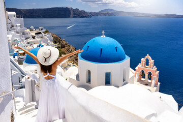A tourist woman in white dress and with outstretched arms enjoys the beautiful view over the village of Oia, Santorini island, Greece, during summer travel time