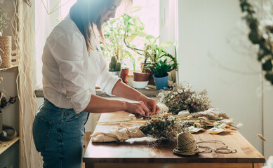 Beautiful Florist Arranging Bouquet of a Dry Flowers at her Workshop.
Side view of young talented woman cutting with scissors dried plants and flowers arrangement in her flower shop. - obrazy, fototapety, plakaty