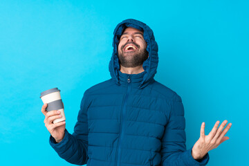 Man wearing winter jacket and holding a takeaway coffee over isolated blue background smiling a lot