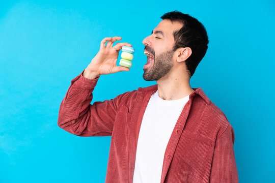 Young caucasian man over isolated blue background holding colorful French macarons and eating it