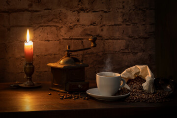 Bodegón con una taza de café un molinillo , un saquito con granos de cafés y todo alumbrado a la luz de una vela y con una pared de ladrillos de fondo .