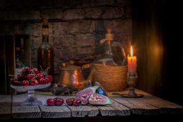 Fototapeta na wymiar Imagen de un bodegón rústico con cerezas , Higos , una garrafa , cafetera , botella de vino y un candelabro con una vela encendída y con una pared de ladrillos de fondo .