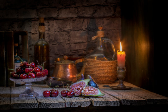 Imagen de un bodegón rústico con cerezas , Higos , una garrafa , cafetera , botella de vino y un candelabro con una vela encendída y con una pared de ladrillos de fondo .