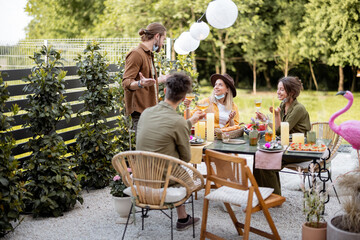 Young friends in face mask having festive dinner during a summertime at the backyard of the country house. Happy to meet each other in real life after pandemic