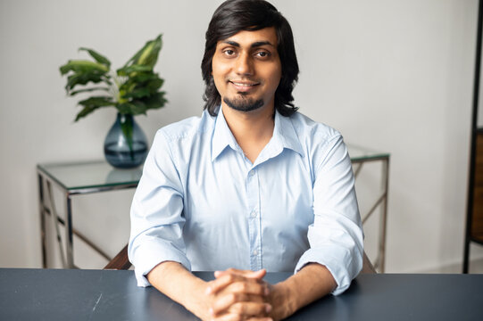 Portrait Of Happy Indian Man Sitting At The Office Desk And Looks At The Camera, Involved In Video Meeting, Webcam View Of Smart Eastern Male In Blue Shirt