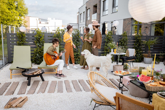 Friends hang out together on a beautiful cottage garden, playing on a guitar, playing with a dog and eating some snacks. Wide view on residential backyards