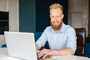 Ambitious redhead bearded male entrepreneur looks at the camera with a friendly toothy smile, young bearded man in shirt sits at the desk and using laptop indoor, male office worker