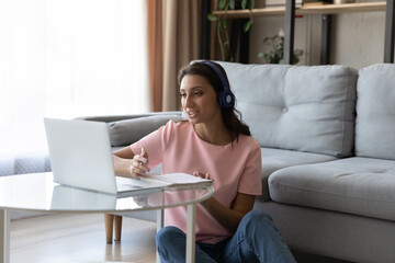 Fototapeta na wymiar Smiling Arabian woman in headphones studying online, taking notes, using laptop at home in living room, motivated young female watching webinar, involved in online course, preparing to exam
