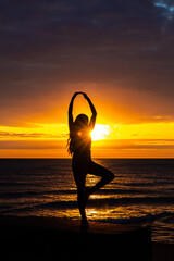 A beautiful African American woman doing a ballerina pose creates sunrise silhouette on Lake Michigan as she looks out across the glistening water with orange and yellow clouds in the sky above.