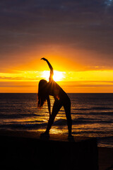 A beautiful sunrise silhouette of an athletic woman stretching her arms and back in front of Lake Michigan water reflecting an incredible bright orange and yellow colored sky.