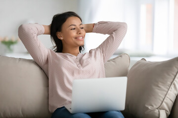 Happy relaxed female student, freelancer, customer relaxing on couch with laptop at home, resting head o hands, looking away, thinking over good future, enjoying work break, breathing fresh air