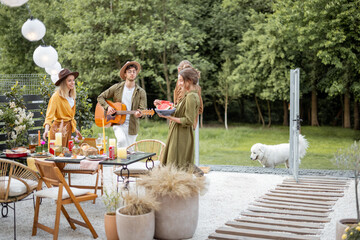 Friends hang out together on a beautiful terrace near the forest, talking and playing with dog and guitar, having great summertime