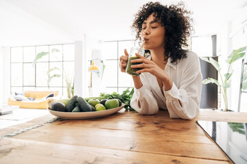 Young african american woman drinking green juice with reusable bamboo straw in loft apartment - obrazy, fototapety, plakaty