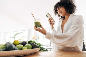 Young african american woman taking photo of green juice and vegetables with cellphone at home. - obrazy, fototapety, plakaty