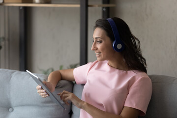 Profile of smiling Arabian woman in headphones using tablet, sitting on couch at home, happy young female looking at device screen, typing, writing message in social network, watching video