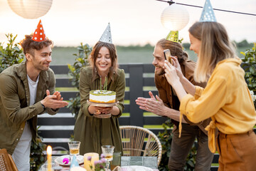 Young adult friends celebrating birthday, having fun blowing candles on a cake at backyard of a...