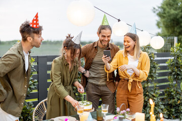 Young adult friends celebrating birthday, having fun blowing candles on a cake at backyard of a...