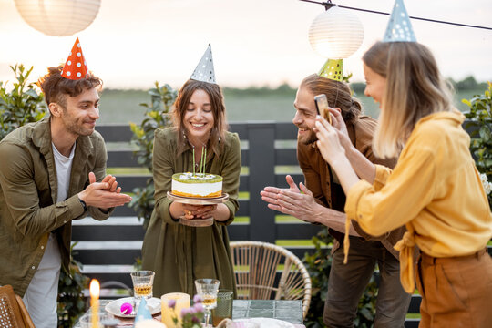 Young adult friends celebrating birthday, having fun blowing candles on a cake at backyard of a country house outdoors