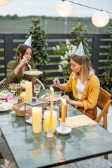Two female friends celebrating birthday alone, sitting together by a festive table at backyard. eating sweet cake