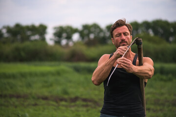 Farmer in sleeveless shirt sharpening scythe in field in morning 