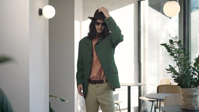 Stylish confident man in sunglasses putting on hat in slow motion standing indoors in 1980s. Portrait of serious fashionable dandy posing in vintage retro cafe. 1990s style and lifestyle