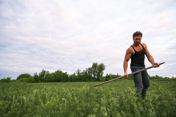 Athletic farmer with scythe standing in field with sky at background in morning 