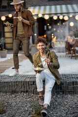 Obraz premium Two male friends eating sweet cakes on a porch of the country house on dusk outdoors. Fun summer time in a country house, male friendship