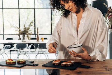 Young african american woman preparing smoked salmon toast for breakfast on wood kitchen counter top